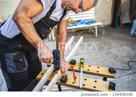 Mature male carpenter using electric drill on a wooden plank outdoors 130498586