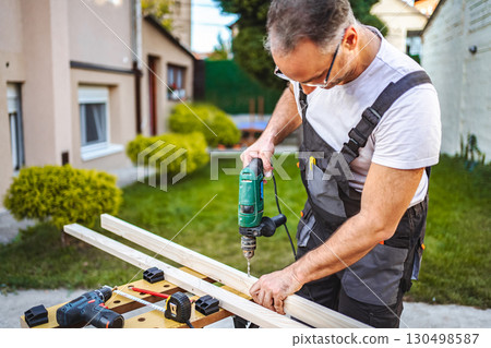 Mature male carpenter using electric drill on a wooden plank outdoors 130498587