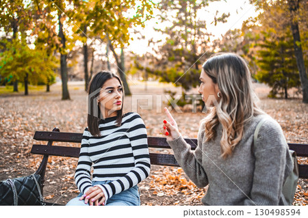 Two women are sitting on a park bench and are angrily pointing fingers at each other 130498594