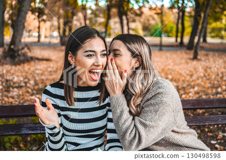 Two women whisper a secret in each other's ear and share a laugh Two women whisper a secret in each other's ear and share a laugh 130498598