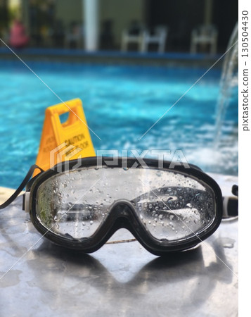 Swimming goggles with water droplets rest on a wet surface by a pool. A yellow CAUTION WET FLOOR sign is blurred in the background, suggesting summer recreation and safety. Swimming goggles with water droplets rest on a wet surface by a pool. A yellow CAUTION WET FLOOR sign is blurred in the background, suggesting summer recreation and safety. 130504430