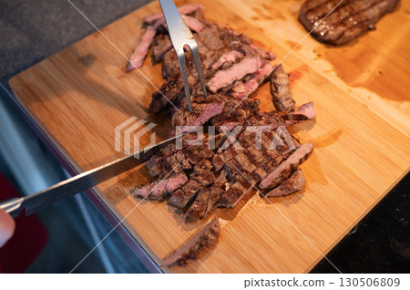 Grilled steak slices on wooden cutting board being cut with knife and fork Grilled steak slices on wooden cutting board being cut with knife and fork 130506809