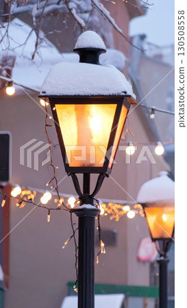 Snow Covered Black Street Lamp Glowing Warmly at Dusk in Winter City Landscape 130508558