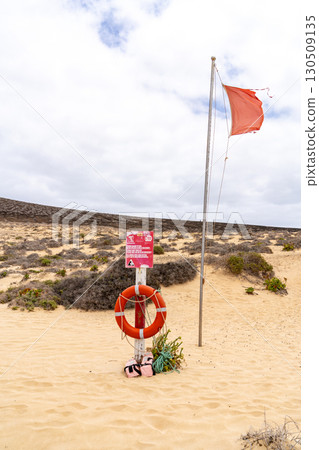 A red flag on a beach indicates dangerous conditions, next to life-saving equipment in a sandy coastal environment. A red flag on a beach indicates dangerous conditions, next to life-saving equipment in a sandy coastal environment. 130509135