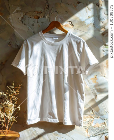 White T Shirt Hanging on Wooden Hanger Against Textured Beige Wall with Floral Decor Sunlight 130509575
