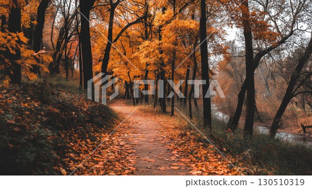 A winding path covered in vibrant autumn leaves leads through a peaceful forest beside a gentle river 130510319