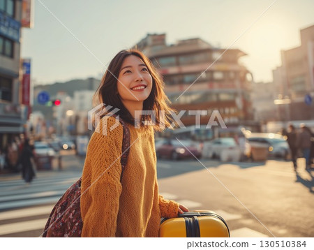 Smiling Korean woman looks up with a yellow suitcase on a bright city street 130510384