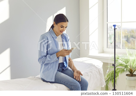 Young smiling woman using smartphone while receiving IV drip infusion and vitamin therapy. 130511604