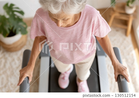 Top view of a senior woman walking on a treadmill at home, wearing a pink shirt and sneakers, staying active and focused on fitness in a cozy setting Top view of a senior woman walking on a treadmill at home, wearing a pink shirt and sneakers, staying active and focused on fitness in a cozy setting 130512154