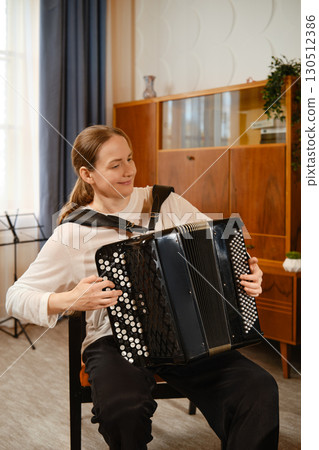 A middle-aged woman plays the accordion in a cozy living room, showcasing her musical talent during daylight 130512386