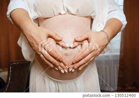 Expecting parents create a heart shape over baby bump in cozy indoor setting Expecting parents create a heart shape over baby bump in cozy indoor setting 130512400