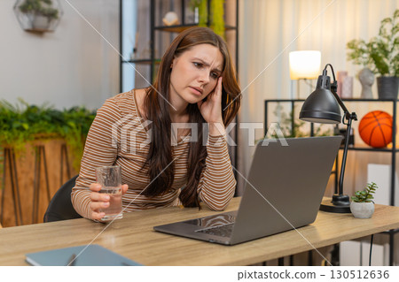 Young woman rubs temples after long laptop work drinks water squints from fatigue and breathes slow Young woman rubs temples after long laptop work drinks water squints from fatigue and breathes slow 130512636
