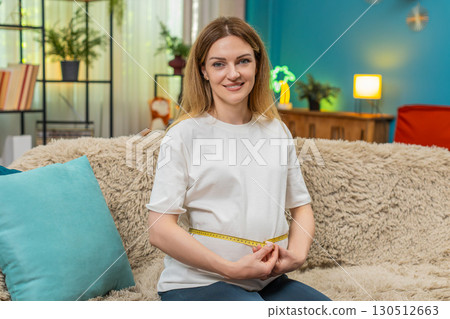 Pregnant woman measures belly with tape checks result smiles happy with nice growth on sofa at home 130512663