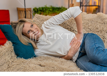 Adult woman holding stomach at home, expressing abdominal pain and discomfort while lying on sofa 130512838