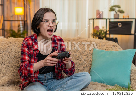 Woman playing video game with gamepad at home, laughing and reacting energetically with fun gestures 130512839