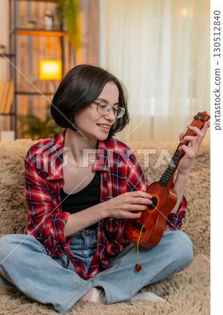 Young woman playing ukulele at home, focused on learning chord and smiling with satisfaction and joy Young woman playing ukulele at home, focused on learning chord and smiling with satisfaction and joy 130512840