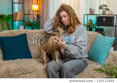 Young woman reacting to bad news on smartphone with pug dog on home sofa, emotional and serious 130512848