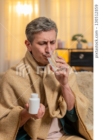 Young man takes pill with tired expression while holding head in pain from headache or migraine 130512859