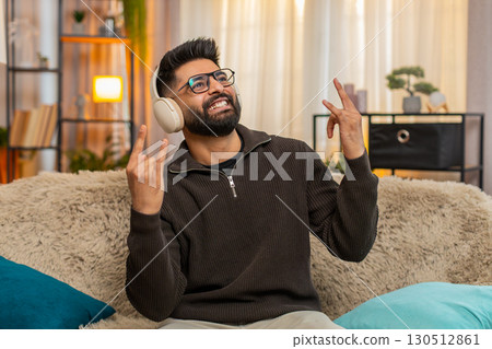 Indian man smiling listens to energetic music in wireless headphones, shows rock-n-roll gesture Indian man smiling listens to energetic music in wireless headphones, shows rock-n-roll gesture 130512861