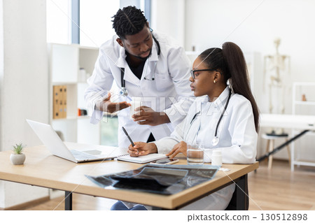 Two medical professionals in white coats discuss and plan patient treatment strategy using laptop in bright modern office. Focus on teamwork, research, patient care concepts. 130512898