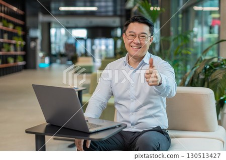 Happy young businessman working on laptop looking at camera showing thumbs up in modern office lobby Happy young businessman working on laptop looking at camera showing thumbs up in modern office lobby 130513427