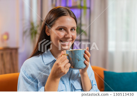 Attractive smiling young woman girl drinking a cup of coffee or herbal tea sitting on home couch Attractive smiling young woman girl drinking a cup of coffee or herbal tea sitting on home couch 130513430