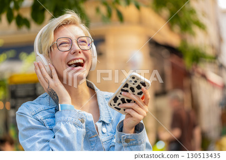 Overjoyed blonde woman in wireless headphones choosing, listening music holding smartphone on street 130513435