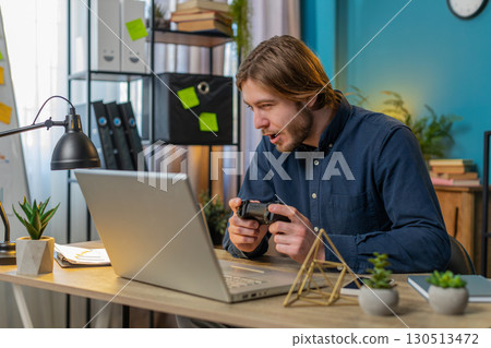 Happy freelancer business man playing video games on laptop holding controller at home office 130513472