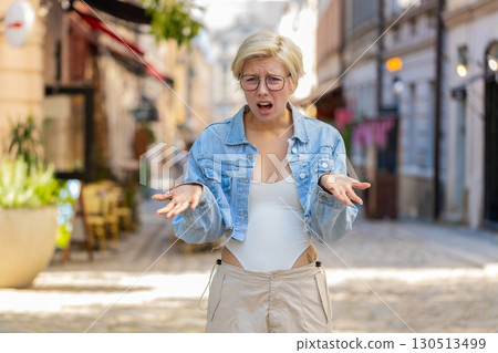 Doubtful sad frustrated young Caucasian woman shrugging shoulders dont know answer on city street 130513499