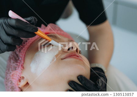 professional cosmetologist applies a creamy face mask on yiung woman face using a brush professional cosmetologist applies a creamy face mask on yiung woman face using a brush 130513857