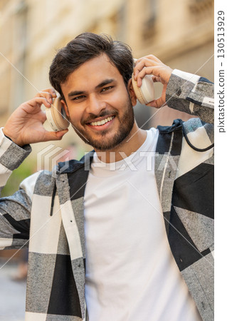 Happy Arabian man taking off wireless headphones and looking at camera smiling in city street Happy Arabian man taking off wireless headphones and looking at camera smiling in city street 130513929