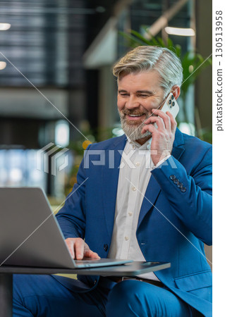 Mature businessman in suit answering to client on mobile phone call, using laptop in office lobby 130513958