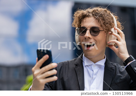 Happy joyful businessman listens to music on wireless headphones while walking on street near office Happy joyful businessman listens to music on wireless headphones while walking on street near office 130513966