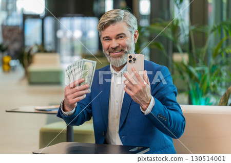 Successful businessman holding smartphone and counting dollar bill money fan sitting in office lobby Successful businessman holding smartphone and counting dollar bill money fan sitting in office lobby 130514001