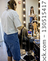 In a trendy salon, a skilled stylist focuses on intricately braiding a client's long hair. A second client relaxes as she prepares for her hairstyling session. 130515417
