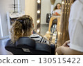 In a cozy salon, a stylist skillfully braids a young girl's hair, while she relaxes on a chair, engrossed in her phone, highlighting a blend of creativity and relaxation. 130515422