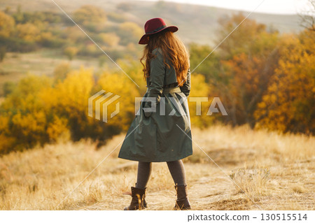 Beautiful stylish girl walks in the autumn park. The girl is dressed in a green coat and a red hat. 130515514
