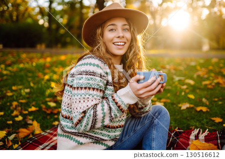 Beautiful woman in stylish clothes enjoys hot drink in hat in clearing among yellow leaves. 130516612