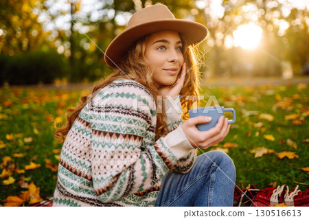 Beautiful woman in stylish clothes enjoys hot drink in hat in clearing among yellow leaves. 130516613