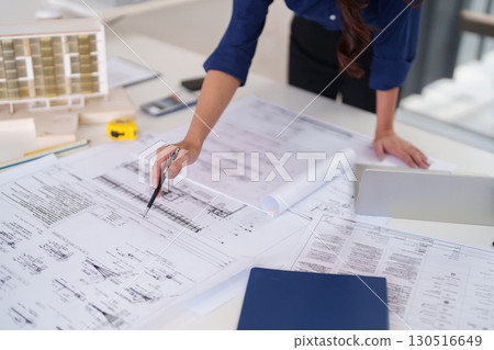 Adult Asian woman hand examines detailed architectural blueprints design plans for construction project planning development in professional office studio 130516649