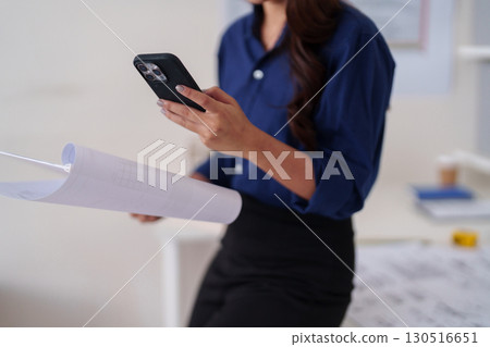 Professional Asian adult architect woman reviews construction plans blueprints designs using smartphone technology manages project operations office workspace 130516651
