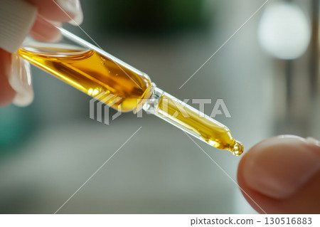 Close up of gloved hand holding glass dropper, releasing a golden viscous liquid drop. Natural plant extract or cosmetic oil being precisely applied. 130516883