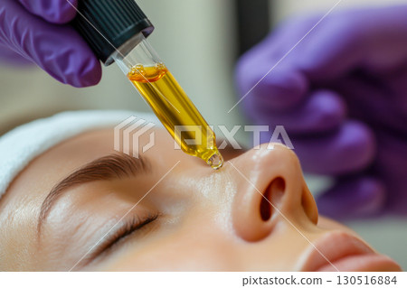 Professional hand in purple glove applies golden facial serum with dropper onto woman's face. Close up of nourishing skincare treatment. 130516884