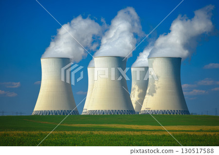 Multiple large industrial cooling towers release white steam against a bright blue sky. A vibrant green field sits below. Power plant energy production. 130517588