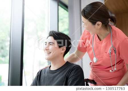 Close-up of a medical worker and a patient in a wheelchair with beautiful fresh greenery by the window. Image of nursing. Close-up of a medical worker and a patient in a wheelchair with beautiful fresh greenery by the window. Image of nursing. 130517763