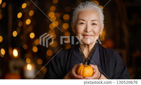 An elderly woman in traditional Japanese clothing smiles indoors with a pumpkin in her hand 130520676