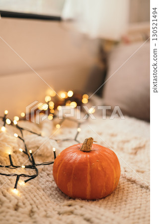 Small pumpkin on cozy knitted blanket with warm fairy lights, autumn home decoration. Small pumpkin on cozy knitted blanket with warm fairy lights, autumn home decoration. 130521494