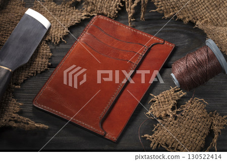 Making leather wallets. On a wooden table there is a leather wallet, a spool of thread and a knife. Making leather wallets. On a wooden table there is a leather wallet, a spool of thread and a knife. 130522414