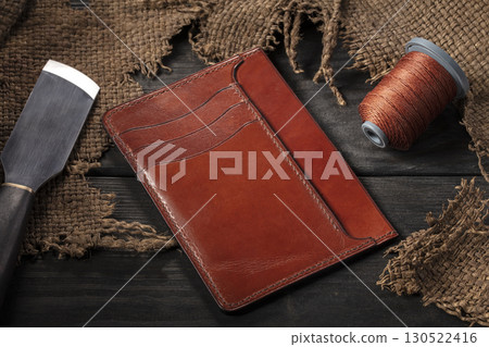 Making leather wallets. On a wooden table there is a leather wallet, a spool of thread and a knife. Making leather wallets. On a wooden table there is a leather wallet, a spool of thread and a knife. 130522416