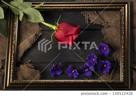 In a beautiful frame, pieces of canvas, a red rose and violets on a wooden background. Decor and decoration. In a beautiful frame, pieces of canvas, a red rose and violets on a wooden background. Decor and decoration. 130522426